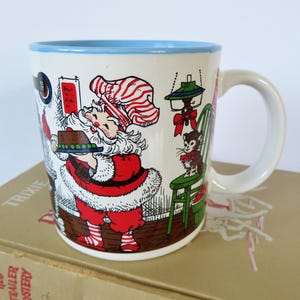 May include: A white ceramic mug with a light blue rim and handle, featuring a colorful illustration of Santa Claus wearing a red and white striped hat, holding a small cake, standing next to a green chair with a small cat sitting on it.