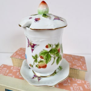 May include: A white ceramic jam jar with a lid, saucer, and gold trim. The jar is decorated with strawberries, butterflies, and insects. The lid has a strawberry-shaped handle. The jar sits on a square saucer.