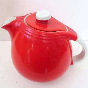 Puede incluir: Una tetera de cerámica roja vibrante con asa y boquilla blancas. La tapa es roja con un pomo blanco. La tetera tiene un cuerpo redondeado y una boquilla curva.