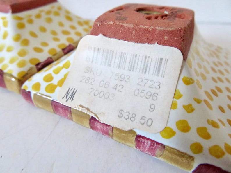 Peut inclure: Gros plan sur deux talons de chaussures d&eacute;coratifs en c&eacute;ramique. Les talons ont une base blanche avec des pois jaunes et une bordure ray&eacute;e rouge et or. Le talon est de couleur brun rouge&acirc;tre. Une &eacute;tiquette de prix est attach&eacute;e.