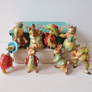 May include: A collection of ceramic figurines depicting anthropomorphic animals. The figurines include a cat, rabbit, mouse, frog, and goose, each dressed in detailed clothing. The figures are arranged on a light-colored surface, suggesting a decorative or collectible theme.