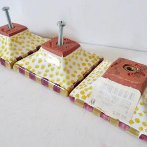 Peut inclure: Ensemble de trois boutons de meuble d&eacute;coratifs. Chaque bouton pr&eacute;sente une base carr&eacute;e brun rouge&acirc;tre avec une vis, sur une surface blanche parsem&eacute;e de points jaunes. Les bords sont orn&eacute;s d'un motif ray&eacute; or et bordeaux.