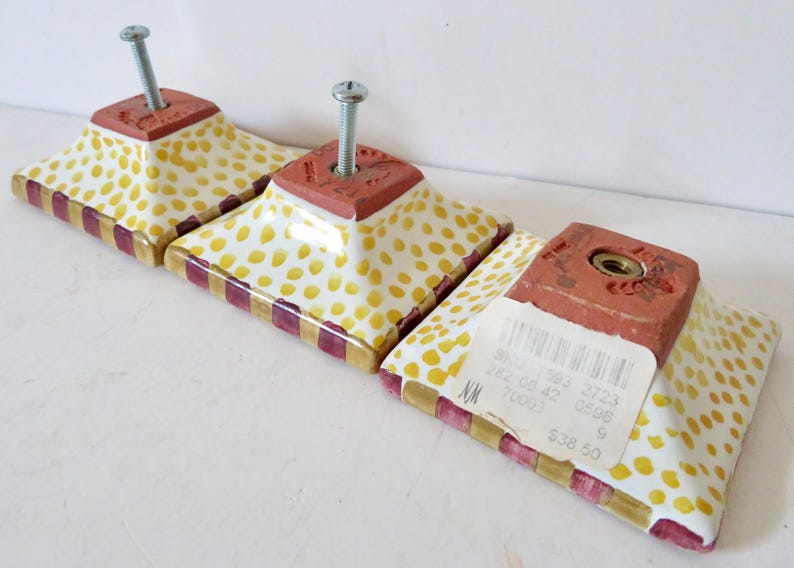 Peut inclure: Ensemble de trois boutons de meuble d&eacute;coratifs. Chaque bouton pr&eacute;sente une base carr&eacute;e brun rouge&acirc;tre avec une vis, sur une surface blanche parsem&eacute;e de points jaunes. Les bords sont orn&eacute;s d'un motif ray&eacute; or et bordeaux.