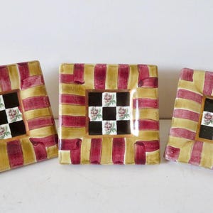 Peut inclure: Trois assiettes d&eacute;coratives carr&eacute;es. Chaque assiette a une bordure ray&eacute;e or et bordeaux. Le centre de chaque assiette pr&eacute;sente un motif &agrave; carreaux noir et blanc avec des accents floraux. Les assiettes sont dispos&eacute;es sur une surface blanche.