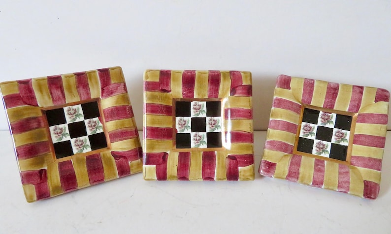 Peut inclure: Trois assiettes d&eacute;coratives carr&eacute;es. Chaque assiette a une bordure ray&eacute;e or et bordeaux. Le centre de chaque assiette pr&eacute;sente un motif &agrave; carreaux noir et blanc avec des accents floraux. Les assiettes sont dispos&eacute;es sur une surface blanche.