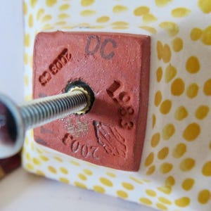 Peut inclure: Gros plan sur un carreau de c&eacute;ramique carr&eacute; avec une vis ins&eacute;r&eacute;e. Le carreau est blanc avec des pois jaunes. Un carr&eacute; rouge avec le texte "DC 1683" et "2003 CS" est estampill&eacute; sur le carreau.