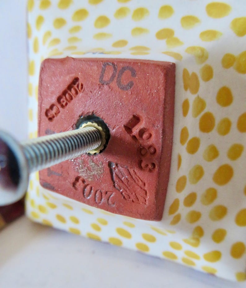 Peut inclure: Gros plan sur un carreau de c&eacute;ramique carr&eacute; avec une vis ins&eacute;r&eacute;e. Le carreau est blanc avec des pois jaunes. Un carr&eacute; rouge avec le texte "DC 1683" et "2003 CS" est estampill&eacute; sur le carreau.