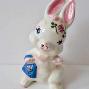 May include: A ceramic bunny figurine with a white base, pink accents, and blue eyes. The bunny holds a blue purse with a floral design and the initials "M.B." The ears and paws are pink, and the bunny has a floral design on its head.