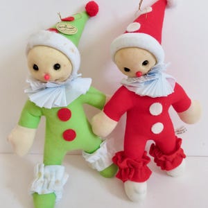 May include: Two vintage clown dolls. One is green with red accents, the other is red with white accents. Both have white ruffled collars, hats, and cuffs. They have small black eyes and red noses. The dolls are made of felt and have a tag attached.