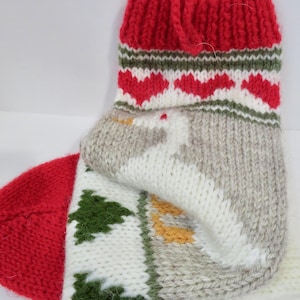 May include: A hand-knitted Christmas stocking with a red cuff and toe. The cuff features red hearts and green stripes. The body of the stocking has a white background with green Christmas trees and a gray background with a white design.