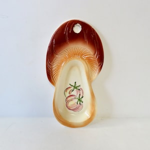 May include: A ceramic spoon rest shaped like a mushroom with a brown and orange glaze. The spoon rest has a hole in the top for hanging and features a painted design of three pink tomatoes.