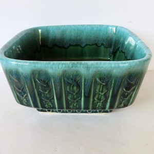 May include: A square ceramic bowl featuring a deep teal and forest green drip glaze finish. The exterior walls are decorated with a repeating vertical relief pattern of stylized floral motifs, while the interior has a smooth, dark green surface.