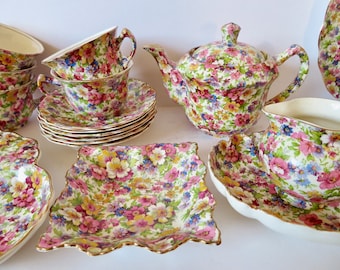 Pode incluir: Um conjunto de chá composto por um bule com tampa, várias chávenas com pires, uma leiteira e pratos quadrados. Cada peça é decorada com um padrão floral denso e colorido de flores cor-de-rosa, amarelas e azuis sobre uma base branca.