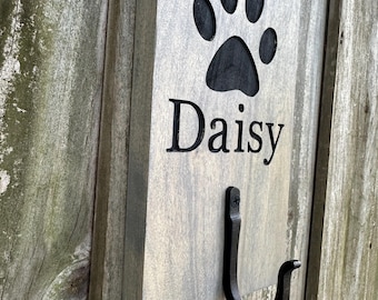 May include: A gray wooden sign with a black paw print and the name "Daisy" engraved on it. A black metal hook is attached to the bottom of the sign.