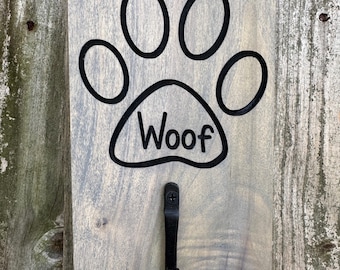 May include: A wooden sign with a black paw print and the word "Woof" carved into it. A black metal hook is attached to the bottom of the sign.
