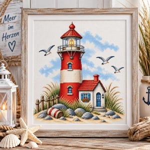 Könnte beinhalten: Eine Kreuzstichstickerei mit einem rot-weißen Leuchtturm und einem kleinen angrenzenden Haus, umgeben von Strandgras, Felsen und drei fliegenden Möwen vor blauem Himmel, gerahmt in einem quadratischen Rahmen aus hellem Holz.