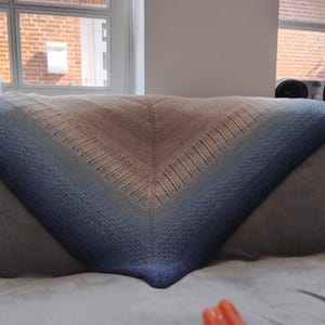 Peut inclure: Un châle tricoté à la main de forme triangulaire, avec un dégradé de couleurs. Le châle passe du beige clair en haut à des nuances de bleu en bas. Le châle est posé sur une surface grise.