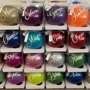 May include: A collection of round, glittery Christmas baubles in various colours, each individually packaged in a clear plastic box with a white ribbon. Every bauble features a unique name written in white cursive script on the front surface.