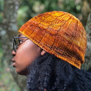 May include: A person wearing a knitted beanie hat with a cable knit pattern in shades of orange and brown. The hat is worn on their head and covers their ears. The person has dark brown hair and is wearing glasses.