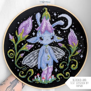 Puede incluir: Un bastidor de madera circular que contiene un diseño de punto de cruz sobre tela negra. La imagen muestra una criatura azul con alas, con un sombrero de flor morada, rodeada de capullos de flores, setas pequeñas y una luna creciente.