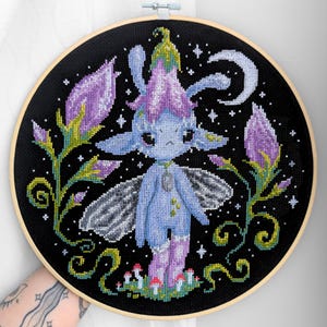Puede incluir: Arte de punto de cruz de un hada azul con detalles florales morados y verdes sobre un fondo negro. El hada tiene alas y está rodeada de estrellas y una luna creciente. La obra está enmarcada en un bastidor de madera.