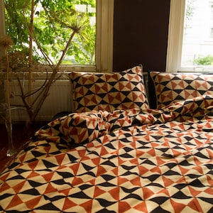 Peut inclure: Ensemble de literie avec un motif géométrique de triangles et de formes incurvées noirs, rouges et crème. L'ensemble comprend une housse de couette et deux taies d'oreiller. La literie est sur un lit près d'une fenêtre.