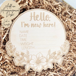 May include: A wooden baby announcement plaque with a floral design. The plaque says "Hello, I'm new here!" and has spaces to fill in the baby's name, date, time, weight, and length.