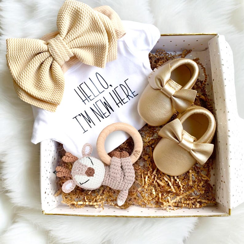 May include: A gift box filled with baby items, including a white onesie with the text "Hello, I'm New Here", a gold bow headband, a pair of gold leather baby shoes, a wooden teething ring, and a crocheted deer toy.
