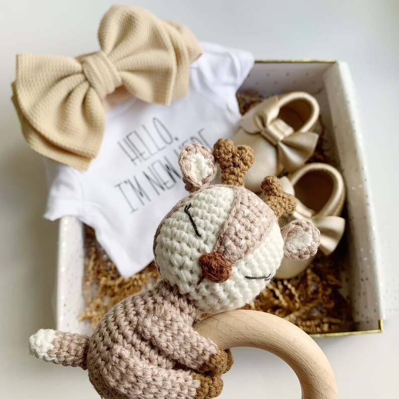 May include: A gift box filled with baby items, including a white onesie with the text "Hello, I'm New Here", a beige bow headband, a crocheted deer rattle, and two pairs of beige baby shoes.