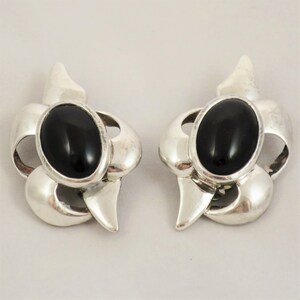 May include: A pair of silver clip-on earrings with black oval-shaped stones set in a floral design.