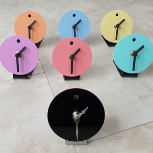May include: Seven small round analogue clocks with black hands and different coloured faces. The clocks are arranged in a circle on a light beige tiled floor. The colours of the clocks are black, pink, light blue, yellow, and turquoise.