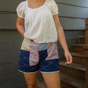 Puede incluir: Una blusa blanca de manga corta con cuello redondo y detalles bordados. Los shorts son patchwork con paneles acolchados azules, lavanda y crema. Los shorts tienen un patrón de nubes.