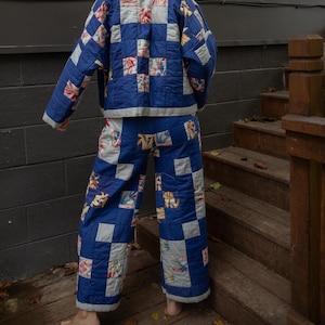 May include: A blue quilted jacket and matching trousers set. The jacket and trousers feature a patchwork design with blue squares and floral print squares. The outfit is worn by a person standing on wooden steps.