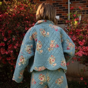 May include: A light blue quilted jacket and pants set with a floral pattern. The jacket has a scalloped hem and the fabric has a diamond pattern. The floral design includes pink, yellow, and cream-colored roses.