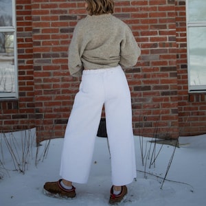 May include: A person wearing a light brown sweater and white pants stands in front of a red brick wall. The person is also wearing brown shoes. The scene is outdoors, with snow on the ground.