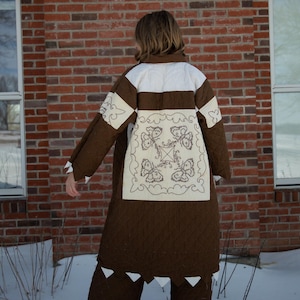 May include: A brown quilted coat with white accents and a decorative square panel on the back featuring embroidered butterfly designs. The coat has a long, flowing design and is paired with matching pants. The background includes a brick wall and snow.