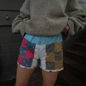 Op de afbeelding: Een persoon draagt een grijze gebreide trui en een patchwork short. De short is gemaakt van verschillende stoffen en heeft een blauw-wit geblokte tailleband.