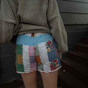 May include: A person wearing a grey sweater and patchwork denim shorts. The shorts are made of different coloured fabrics and have a quilted design.