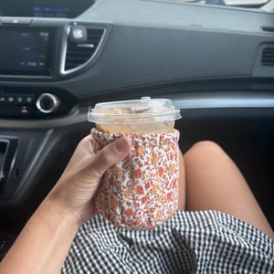 Könnte beinhalten: Eine Tassenmanschette aus Stoff mit einem kleinen Blumenmuster in Orange-, Gelb- und Grüntönen. Die Manschette umschließt einen durchsichtigen Kunststoffbecher, der mit einem Eisgetränk gefüllt ist und einen Kunststoffdeckel sowie einen Strohhalm hat.