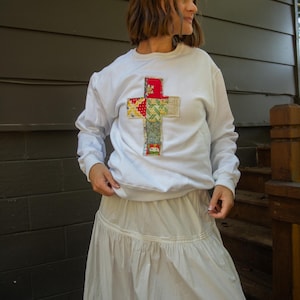 May include: White long-sleeved sweatshirt featuring a patchwork cross design in red, yellow, green, and blue fabrics. The person is wearing a long, white skirt. The background is a dark grey wall and wooden steps.