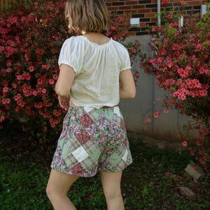 May include: A person wearing a white, short-sleeved blouse and patchwork shorts. The shorts feature floral, plaid, and solid fabric squares in shades of green, burgundy, and white. The person is standing in front of flowering bushes.
