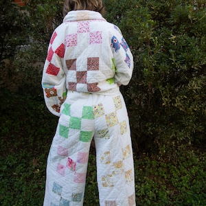 May include: A two-piece quilted outfit consisting of a long-sleeved jacket and wide-leg trousers. The garment is constructed from white fabric featuring a patchwork pattern of small, multi-colored squares in shades of red, pink, green, and blue.