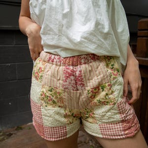 Puede incluir: Pantalones cortos patchwork acolchados con una variedad de estampados florales y a rayas. Los pantalones cortos tienen una cintura elástica y están hechos de telas en tonos rojos, rosas y crema. Parte de una colección de ropa.