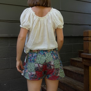 Puede incluir: Una blusa campesina blanca con mangas cortas abullonadas y pantalones cortos patchwork. Los pantalones cortos presentan una mezcla de estampados florales y geométricos en rojo, verde y azul. El atuendo es informal y veraniego.