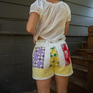 Op de afbeelding: Patchwork shorts met een gele basis en kleurrijke vierkanten stof. De shorts hebben verschillende patronen, waaronder paarse ruiten, tropische bladeren en rode vierkanten. Ze worden gedragen met een witte blouse met korte mouwen.