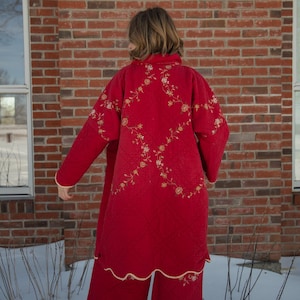 May include: A red quilted robe and matching pants set with floral embroidery. The robe has long sleeves and a collar, and the pants have a wide leg. The set is trimmed with a cream-colored border. The outfit is worn outdoors in front of a brick wall.