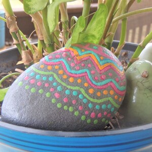 May include: A gray painted rock with a colorful design of pink, green, blue, and orange zigzags and dots. The rock is in a blue pot with green plants and a white ceramic bird.