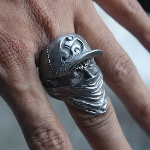 May include: A silver-colored ring featuring a detailed design of a face wearing a cap and a bandana. The ring has intricate detailing, with the face partially obscured by the bandana. The cap has a decorative design on the front.