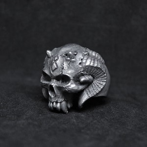 May include: A silver skull ring with horns and intricate details. The ring is made of metal and has a detailed design.