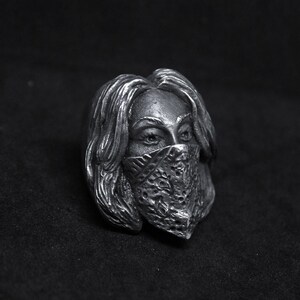 May include: A silver ring with a detailed sculpture of a person's face wearing a bandana. The person's eyes are visible and the bandana has a patterned design.